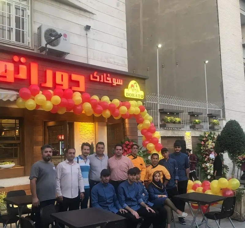 تصویری از فست فود دوراتو، شامل تابلوی روشن "سوخاری دوراتو" و گروهی از پرسنل و کادر در روز افتتاحیه با بادکنک‌های رنگی.