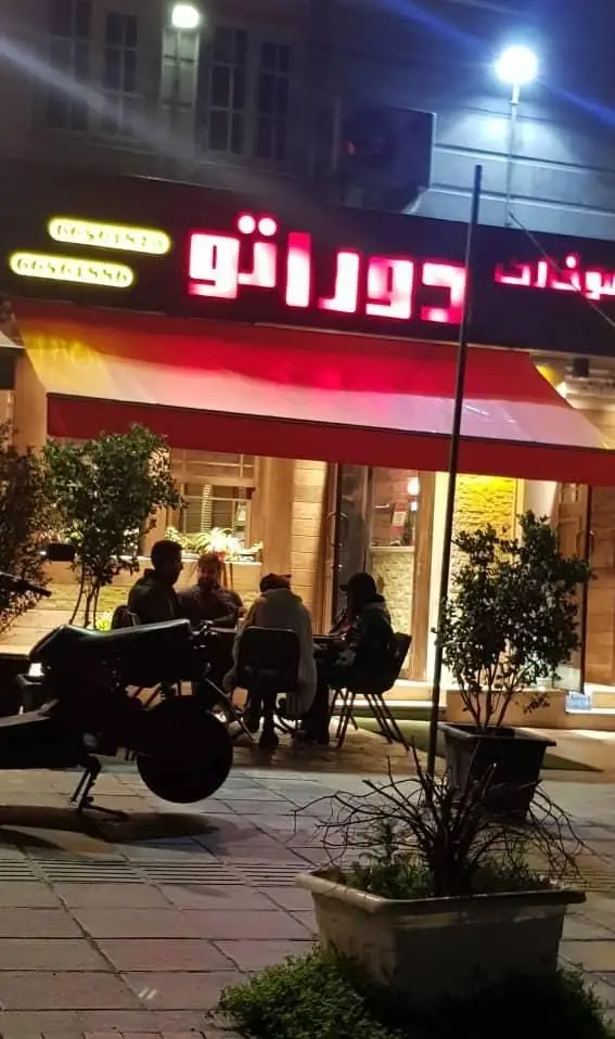 نمای شبانه فست فود دوراتو، تابلوی نئون قرمز روشن، مشتریان در فضای باز نشسته، آویز قرمز و موتور سیکلت پارک شده در ورودی.