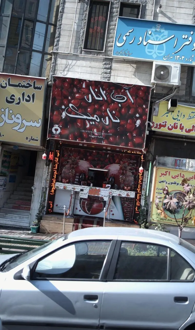 تصویری از کسب و کار آب انار نارمیک در خیابان تهران، شامل غرفه آبمیوه، تابلوهای مشاغل اطراف و خودروی نقره‌ای در حال عبور.