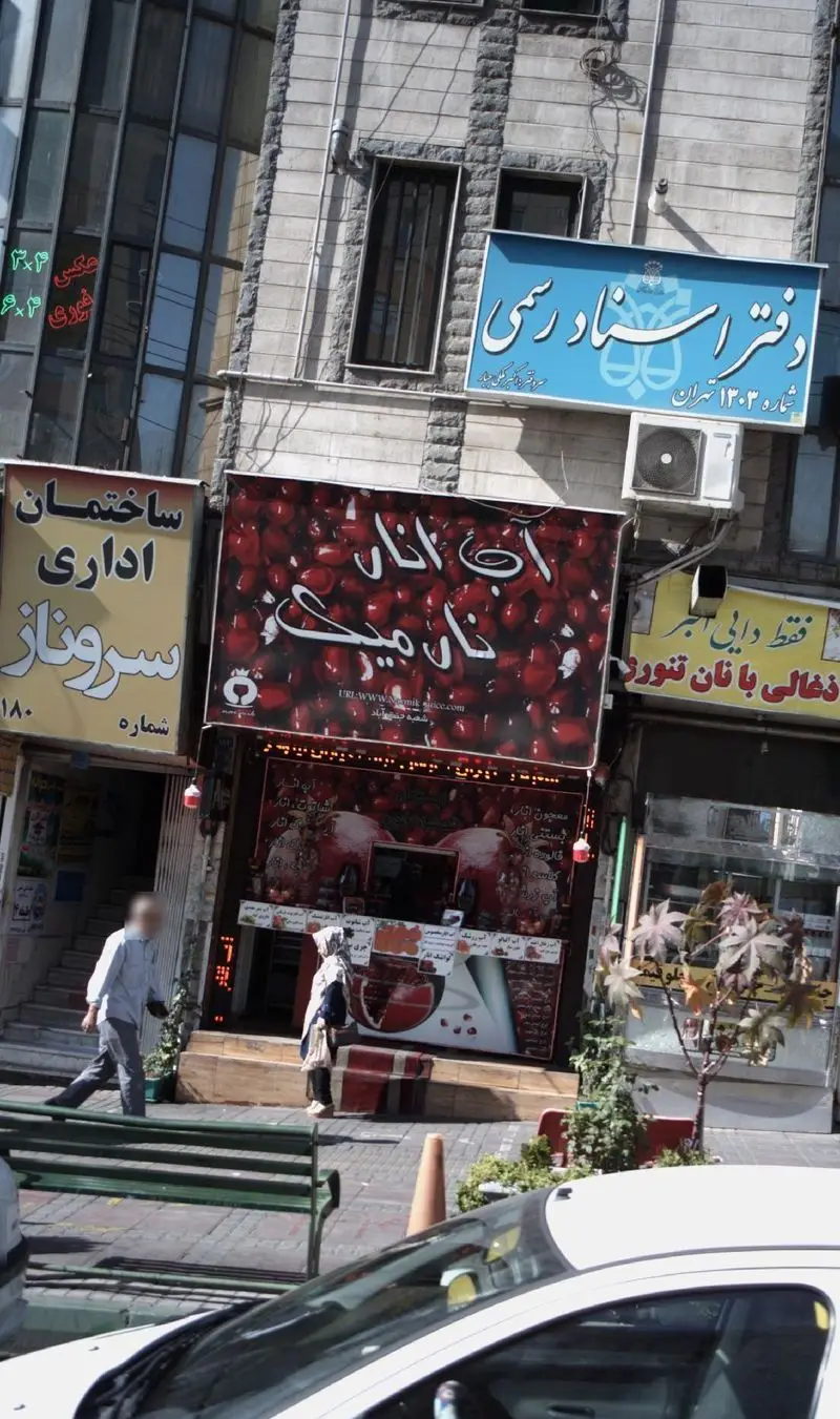 نمایی از فروشگاه آب انار نارمیک در خیابان تهران، با تابلوی قرمز روشن و پس‌زمینه انار، در کنار دفتر اسناد رسمی و ساختمان اداری.