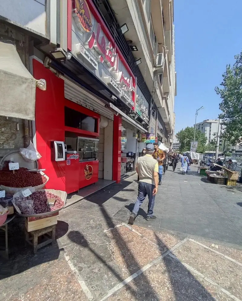 نمای بیرونی رستوران لقمه امیر انفجار با تابلوی قرمز روشن و نام کاربری @Amir_Enfejar. عابران پیاده و بساط میوه‌های خشک در کنار مغازه.