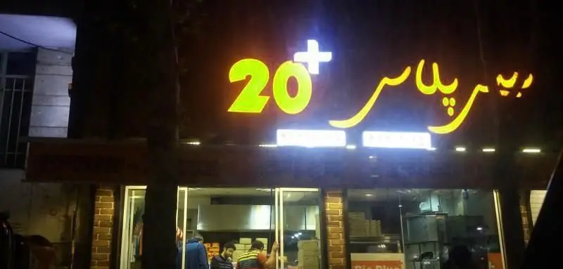 نمای بیرونی روشن فست فود بیس پلاس در شب، با تابلوی نئونی "20+ بیس پلاس" و شیشه‌های مغازه که فعالیت داخلی را نشان می‌دهد.