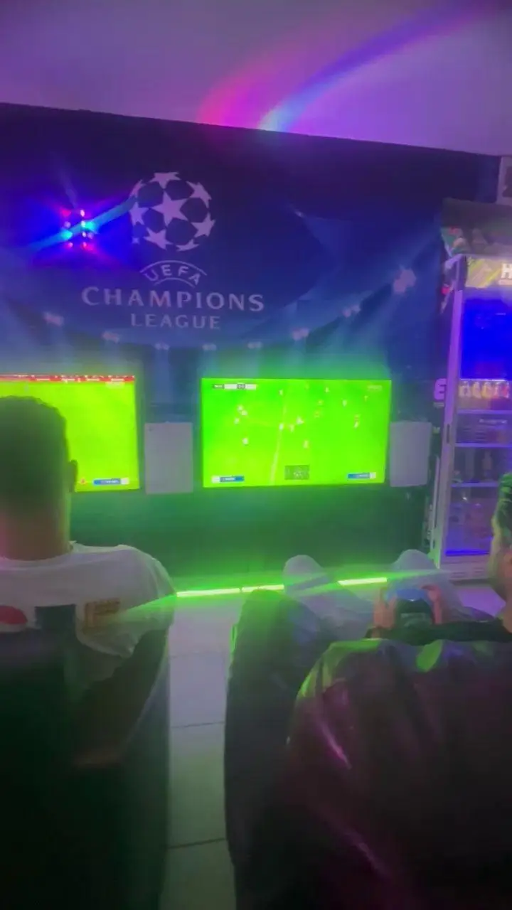 دو گیمر هیجان زده در گیم نت سورنا در حال رقابت فوتبال با پلی استیشن 5 هستند. محیط بازی با نورپردازی جذاب و بنر لیگ قهرمانان اروپا تزیین شده است.