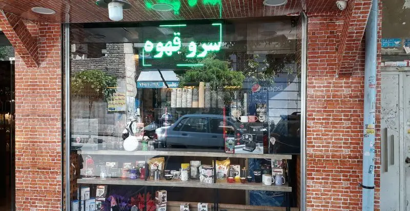 ویترین روشن و چشم‌نواز فروشگاه قهوه کافی لند با تابلوی نئون سبز "سرو قهوه"، عرضه انواع قهوه و لوازم تخصصی دم‌آوری.