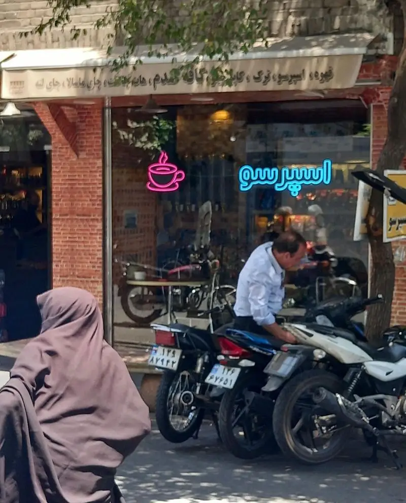 کافه قهوه کافی لند با تابلو نئون اسپرسو و فنجان قهوه، در نمای خیابان با موتورسیکلت‌ها و رهگذران.