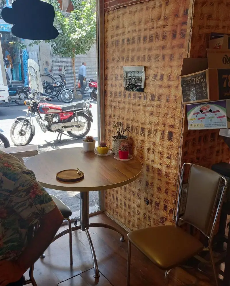 تصویری از محیط داخلی قهوه کافی لند، میزی با گلدان‌های کوچک، دیوار بافت‌دار و چشم‌انداز خیابان شلوغ با موتورسیکلت‌ها.