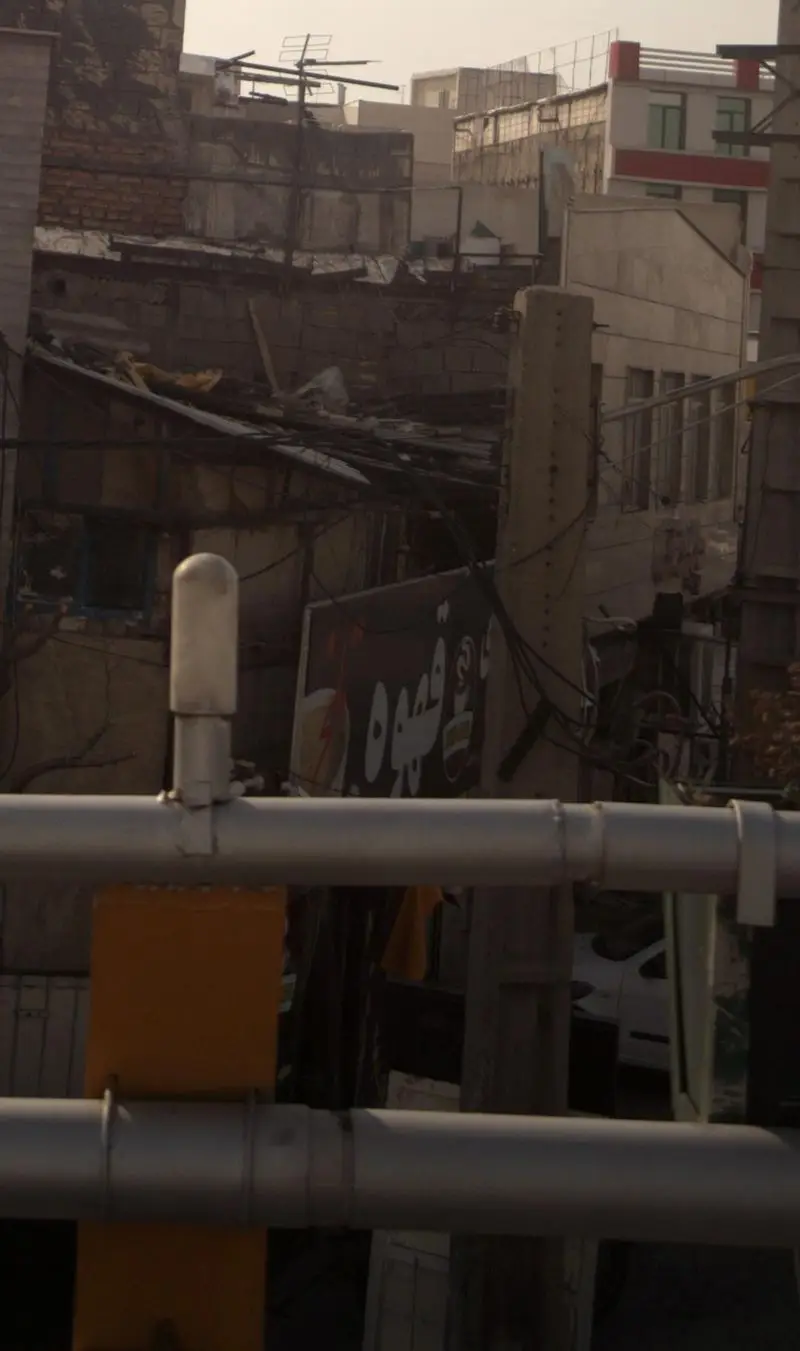 تصویری از نمای شهری با ساختمان‌های متنوع و انبوه سیم‌ها، جایی که تابلوی کافه قهوه تلخ با نماد فنجان قهوه و نوشته "قهوه تلخ" به وضوح دیده می‌شود.