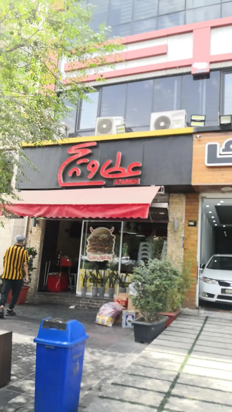 تصویری از نمای بیرونی شعبه رستوران فست فود عطاویچ با تابلوی بزرگ و سایبان قرمز، نمایش پوستر ساندویچ‌های معروف.