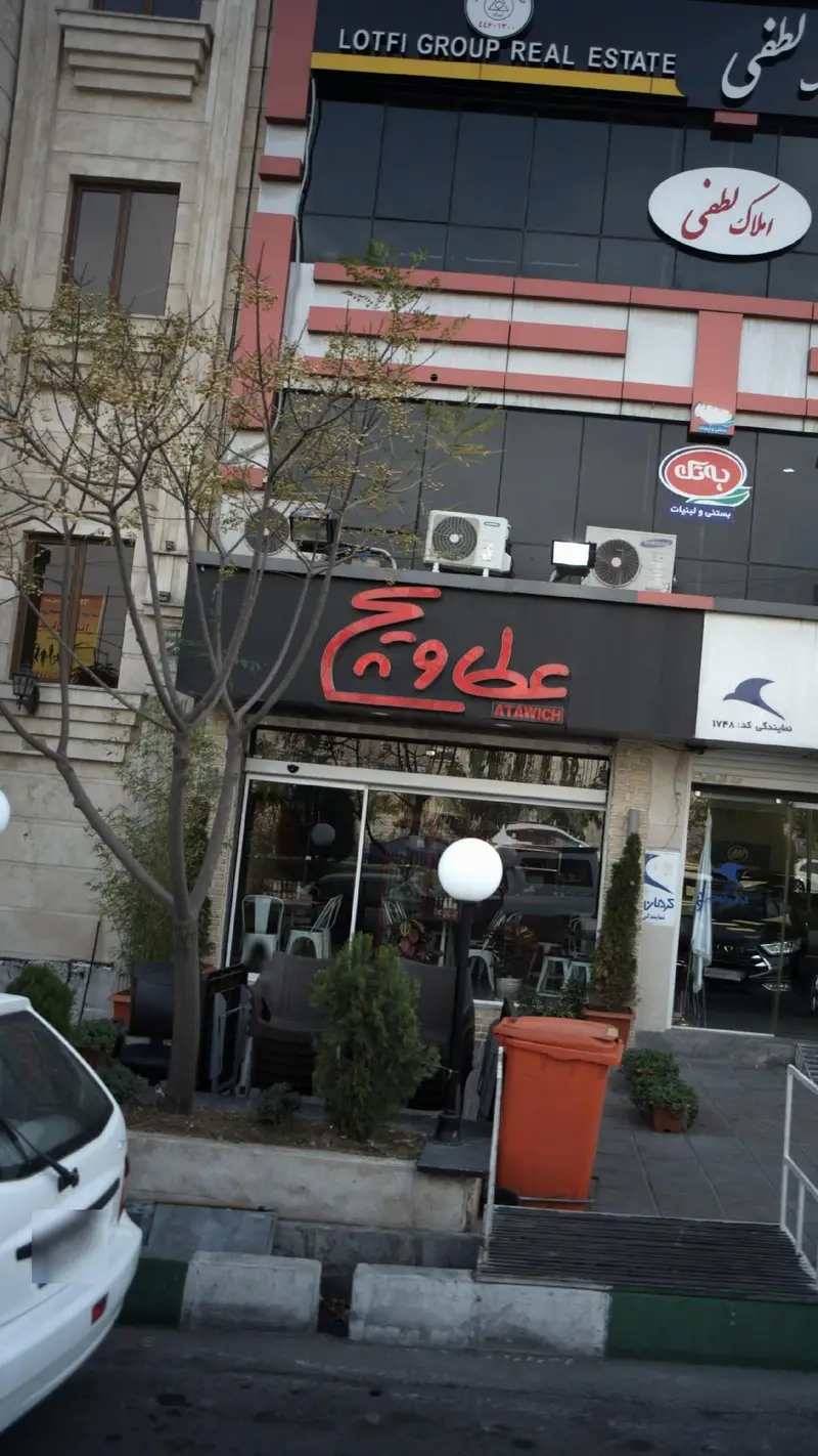 تصویر نمای بیرونی رستوران فست فود عطاویچ، همراه با تابلوی Lotfi Group Real Estate و سایر کسب و کارهای مجاور در خیابان.