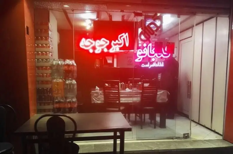 نمای بیرونی رستوران کدبانو با تابلوی نئون قرمز «اکبر جوجه» و «غذا حاضر است»، میز و صندلی‌ها و نوشیدنی‌های آماده سرو.