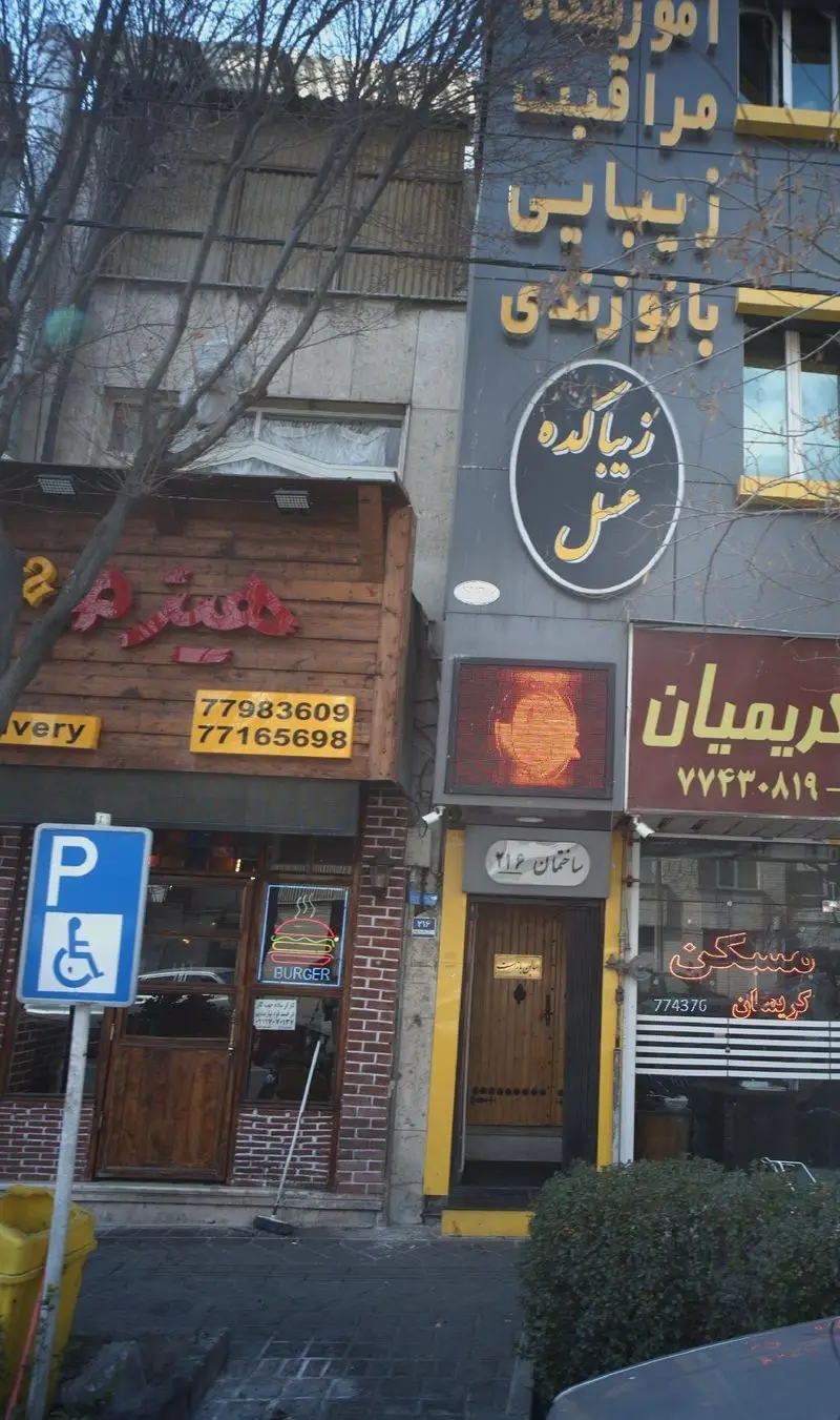 فست فود هیزم، نمای بیرونی با شماره تلفن‌های 77983609 و 77165698 برای دلیوری، تابلو 'BURGER' نئونی و پارکینگ معلولین.
