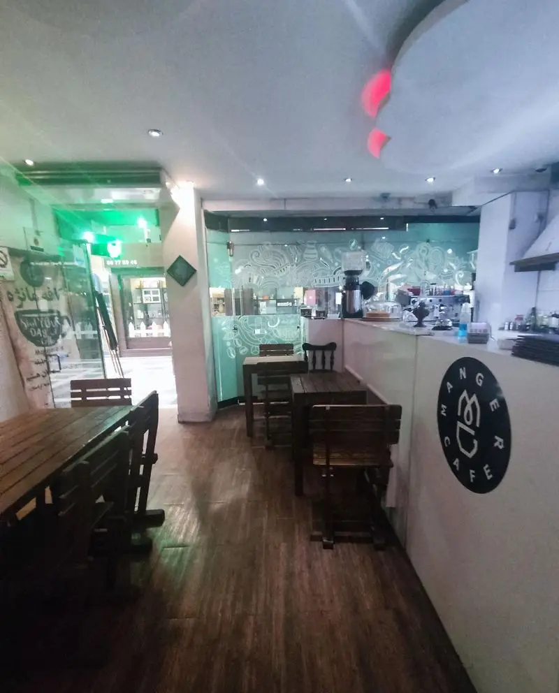 فضای داخلی جذاب و دنج کافه مانژه با دکوراسیون چوبی، میز و صندلی‌های شیک و کانتر مجهز سرو قهوه. لوگو کافه مانژه روی پیشخوان دیده می‌شود.