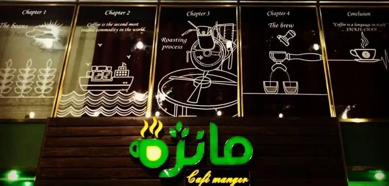 تصویری از داخل کافه مانژه با تابلوی نئون سبز رنگ و نقاشی‌های خطی مراحل قهوه: از دانه، حمل، رست، دم‌آوری تا فنجان نهایی.