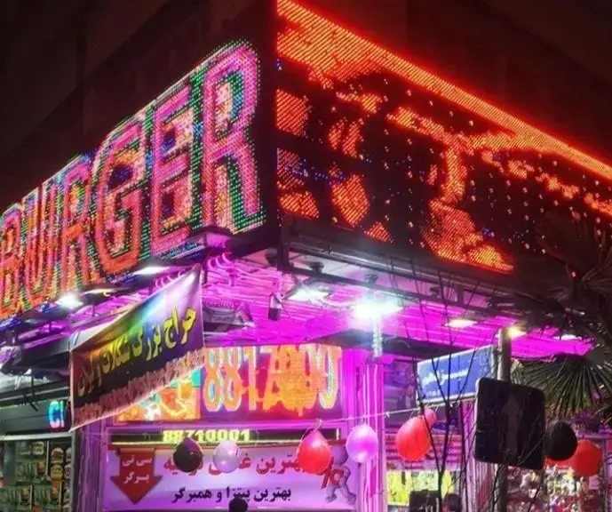 نمای جذاب و پر نور فست فود سیتی برگر در شب با تابلوی LED رنگارنگ "BURGER" و بنر حراج بزرگ پیتزا و انواع برگرهای خوشمزه.