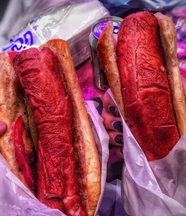 دو ساندویچ هات داگ حجیم و قرمز رنگ از منوی فست فود سیتی برگر که توسط دست ها نگهداری می شوند، با نوشابه و دستمال در پس زمینه، تصویری دلپذیر از غذای آماده.