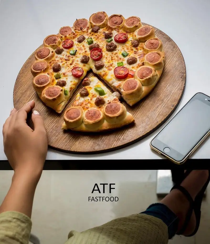 پیتزا فست فود ای تی اف، خوشمزه و خاص با کراست سوسیسی، روی تخته چوبی، تکه‌ای از آن جدا شده و آماده سرو است.