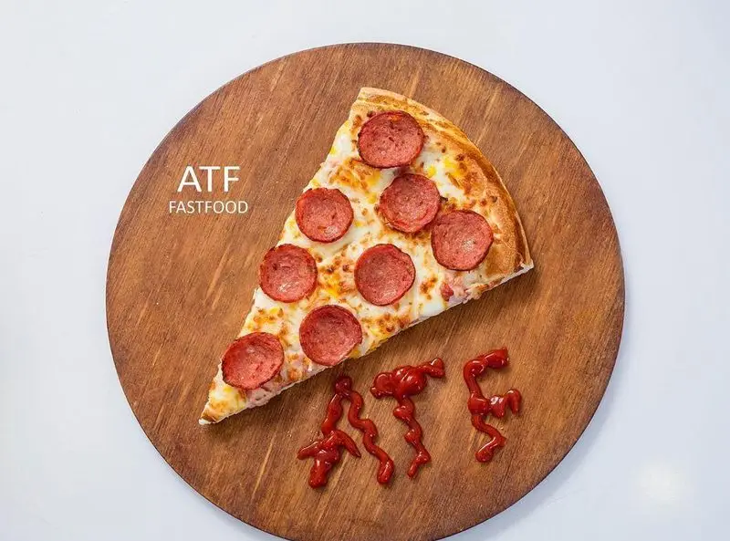 یک برش پیتزا پپرونی داغ و تازه از فست فود ATF روی تخته چوبی، همراه با نام برند ای تی اف و سس کچاپ.