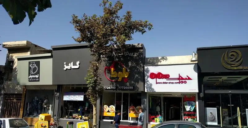 نمای بیرونی فست فود بلوط با تابلوی قرمز رنگ، لوگوی شعله و شماره تلفن های 7726700 و 77726699 در یک خیابان شلوغ.