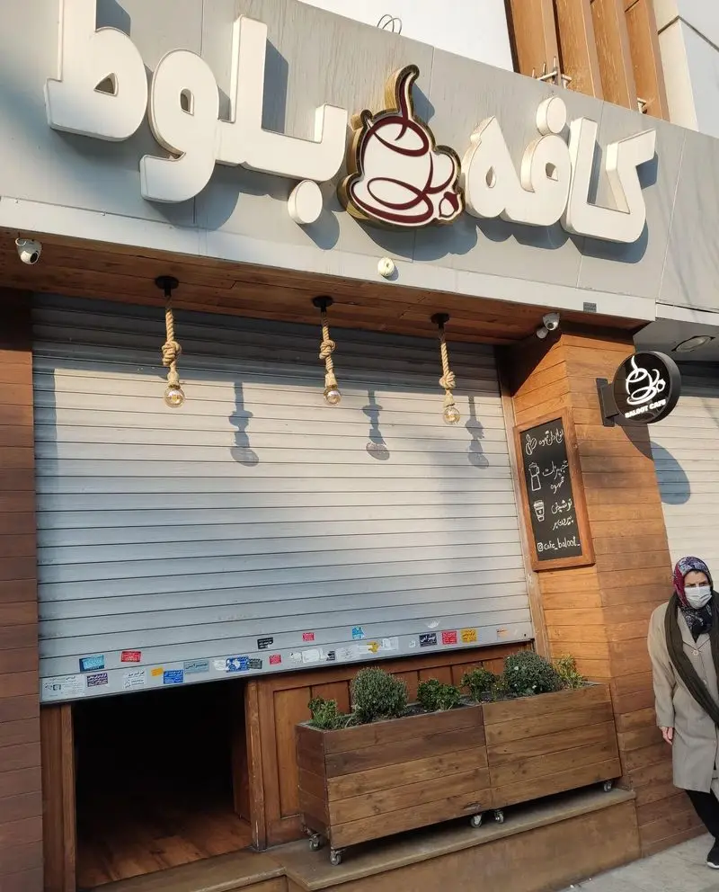 نمای ورودی کافه بلوط با تابلوی روشن و برجسته، لوگوی فنجان قهوه، درب شاتر فلزی بسته و گلدان‌های چوبی تزئینی.