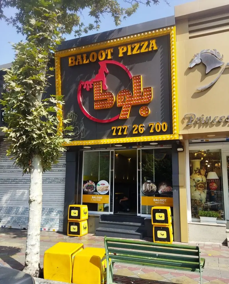 نمای بیرونی فست فود بلوط با تابلوی بزرگ BALOOT PIZZA و شماره تلفن 77726700، شامل ورودی شیشه‌ای و جعبه‌های تحویل غذا.