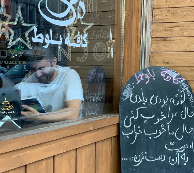 نمایی از فست فود بلوط، مردی جوان در کافه‌ای دنج مشغول خواندن کتاب. تابلو چاک‌بورد با متن فارسی و دکوراسیون چوبی.