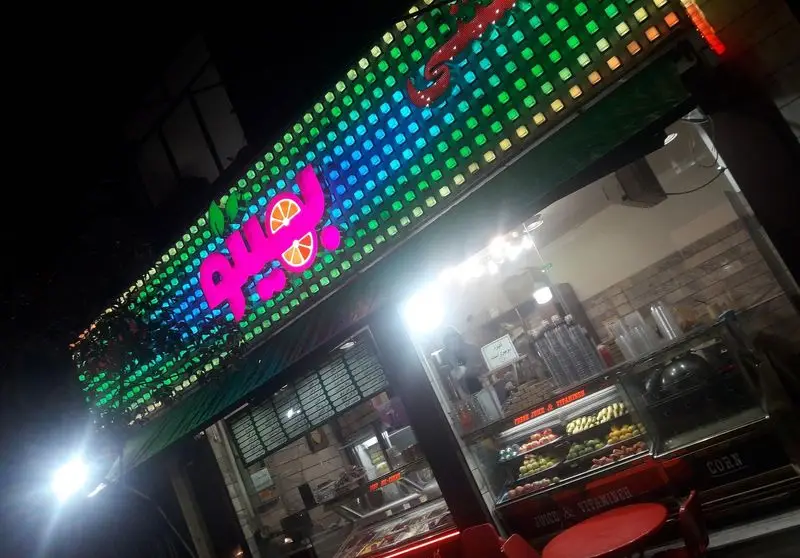 نمای شبانه مغازه آبمیوه و ویتامینه بهیتو با تابلوی نئون رنگارنگ و میوه‌های تازه در ویترین، مکانی جذاب برای نوشیدنی‌های سالم.