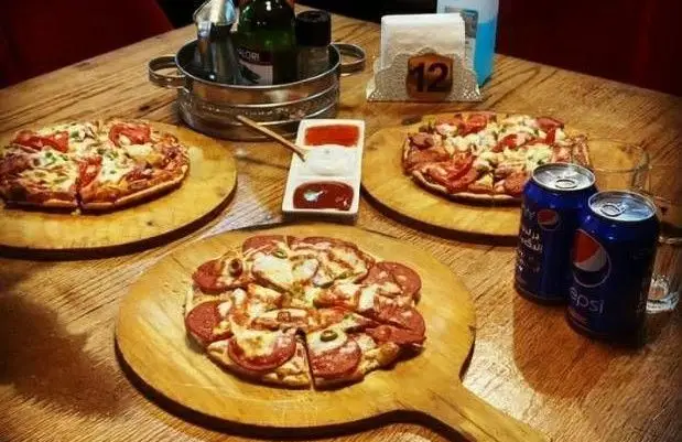میز چوبی کافه راندوو با سه پیتزای خوش طعم، شامل پیتزا پپرونی و سبزیجات، همراه با نوشابه پپسی و سس‌های متنوع برای مشتریان.