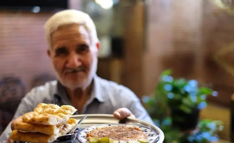 مردی مهربان در سفره خانه سنتی کریم خان، نان داغ تازه و پیش غذای محلی خوش‌طعم را با لبخند به مهمانان ارائه می‌دهد.