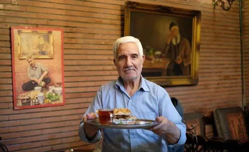 مردی دوست‌داشتنی در سفره خانه سنتی کریم خان در حال ارائه سینی چای و غذای سنتی، تجلی‌گر مهمان‌نوازی و فرهنگ اصیل ایرانی در محیطی دلپذیر.