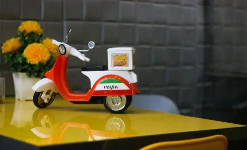 فست فود میلاد: موتور دلیوری وسپا قرمز و سفید روی میز زرد. پیک موتوری آماده برای ارسال سریع غذای آماده و سفارش آنلاین.