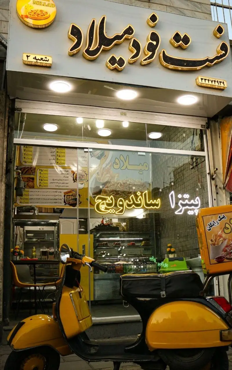 ورودی فست فود میلاد، شعبه ۲، با تابلوی درخشان، منوی پیتزا و ساندویچ و یک اسکوتر زرد رنگ تحویل سفارش.