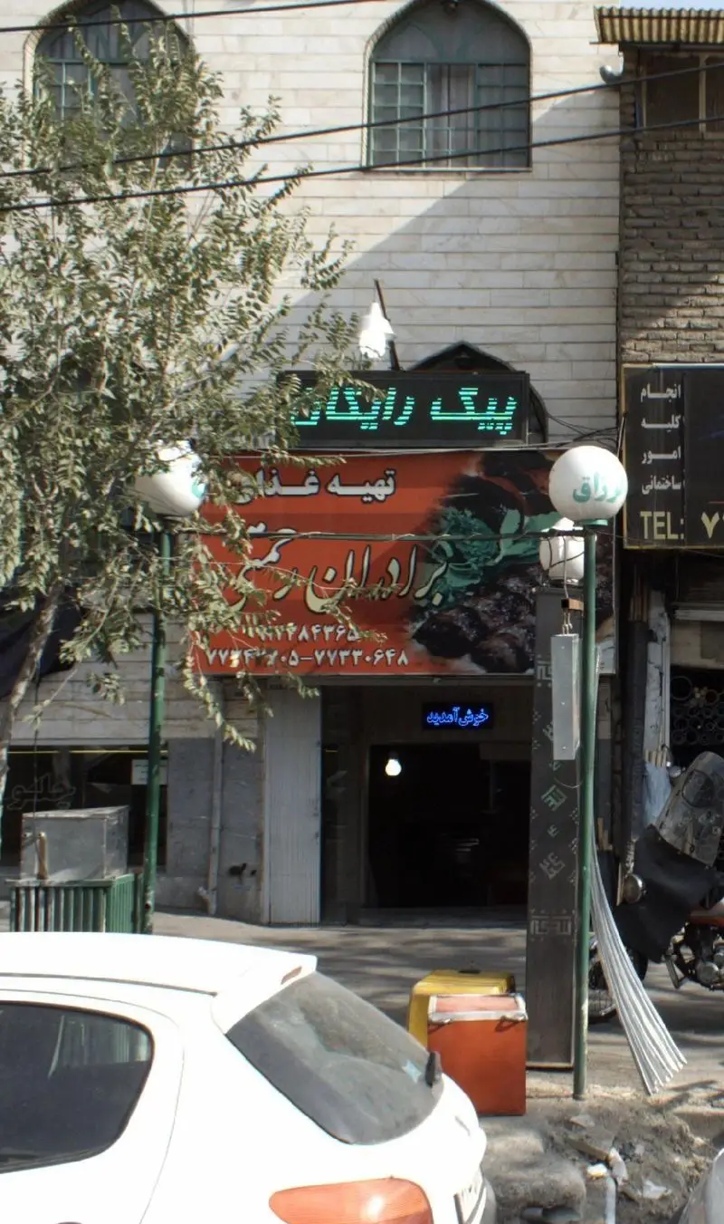 تصویر نمای خیابانی از تهیه غذای برادران رحمتی در تهران با تابلوی پیک رایگان، شماره تماس ۷۷۳۳۰۶۴۸ و ۷۷۳۳۰۶۴۵، عرضه کننده انواع کباب و غذای ایرانی.