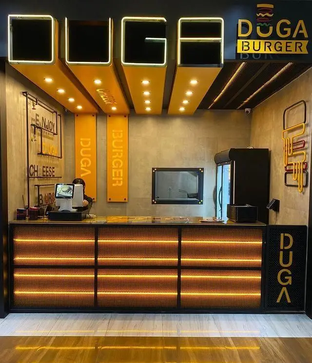 فضای داخلی مدرن و جذاب فست فود دوگا برگر، با کانتر نورانی، دکوراسیون شیک زرد و طوسی و لوگوهای برند روی دیوار و سقف.