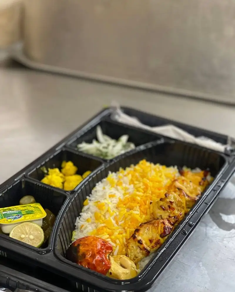 یک پرس غذای کترینگ ته چین ناب شامل کباب جوجه، برنج زعفرانی و سفید، گوجه کبابی، کره، لیمو و مخلفات در بسته بندی بیرون بر