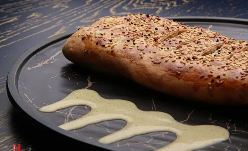 نان داغ و تازه کنجدی فست فود کامنت، برشته و طلایی، روی بشقاب مرمری تیره همراه با سس مخصوص و جذاب.