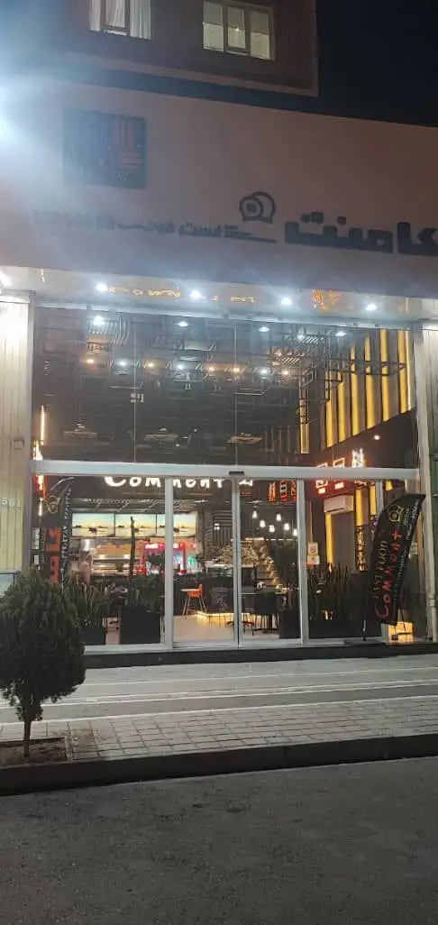 تصویر شبانه از نمای بیرونی فست فود کامنت، یک رستوران مدرن با ویترین شیشه‌ای بزرگ، نورپردازی داخلی روشن و لوگوی برند.