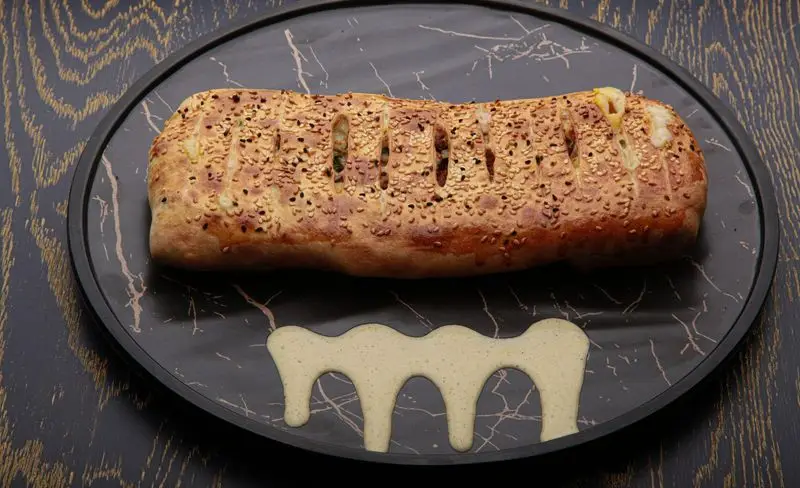 نان شکم پر بزرگ و برشته با کنجد و پنیر، از فست فود کامنت. این غذای خوش‌طعم و طلایی روی بشقاب مرمری تیره همراه با سس مخصوص سرو شده است.