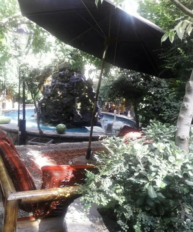 نمای زیبا و آرامش‌بخش رستوران فرنگی آبی با فضای باغ، آبنما، چتر بزرگ و فرش‌های ایرانی در محیطی دلنشین برای صرف غذا.