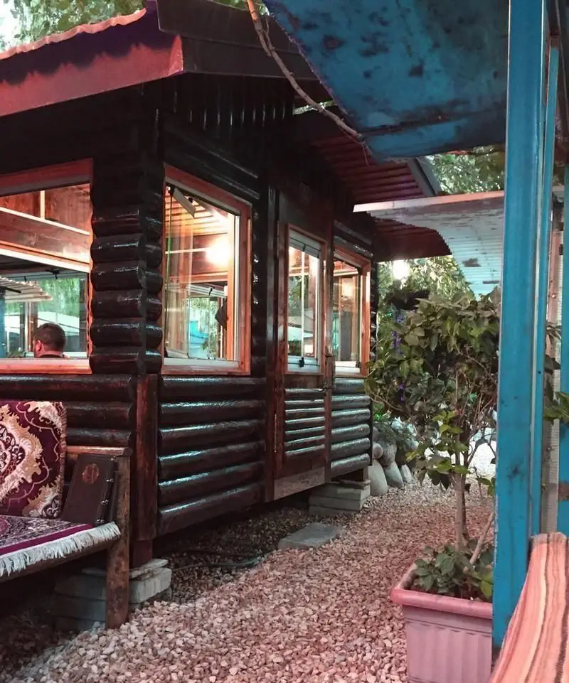 نمای دنج و زیبای رستوران فرنگی آبی با معماری چوبی، پنجره‌های روشن و فضای بیرونی آرامش‌بخش با سنگریزه و گیاهان. مکانی ایده‌آل برای صرف غذا.