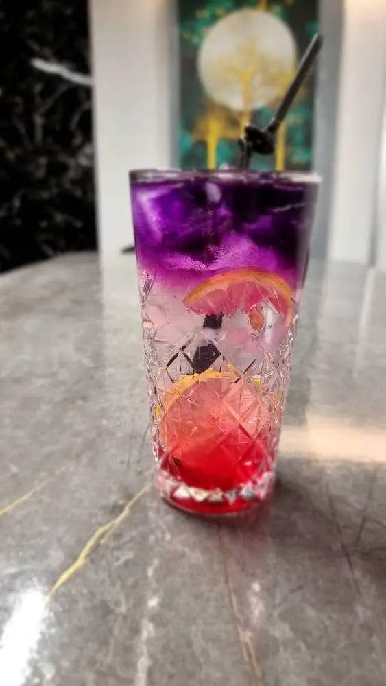 تصویر یک نوشیدنی کوکتل/موکتل لایه ای بنفش و قرمز با لیمو و یخ، نمادی از طعم و زیبایی در کافه رستوران هاید لانژ.
