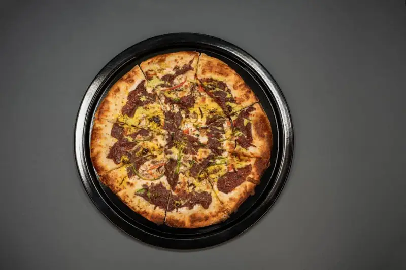 پیتزای مخصوص کافه رستوران هاید لانژ با تاپینگ گوشت، سس ویژه و سبزیجات تازه، برش خورده روی سینی مشکی، آماده سرو.