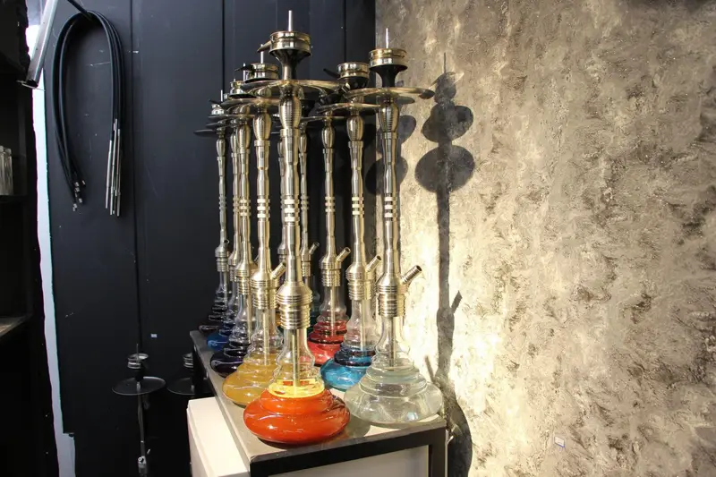 کلکسیونی از قلیان‌های شیک و باکیفیت با پایه‌های رنگارنگ و بدنه طلایی، چیده شده در فضایی دنج و مدرن کافه رستوران هاید لانژ برای تجربه خاص.