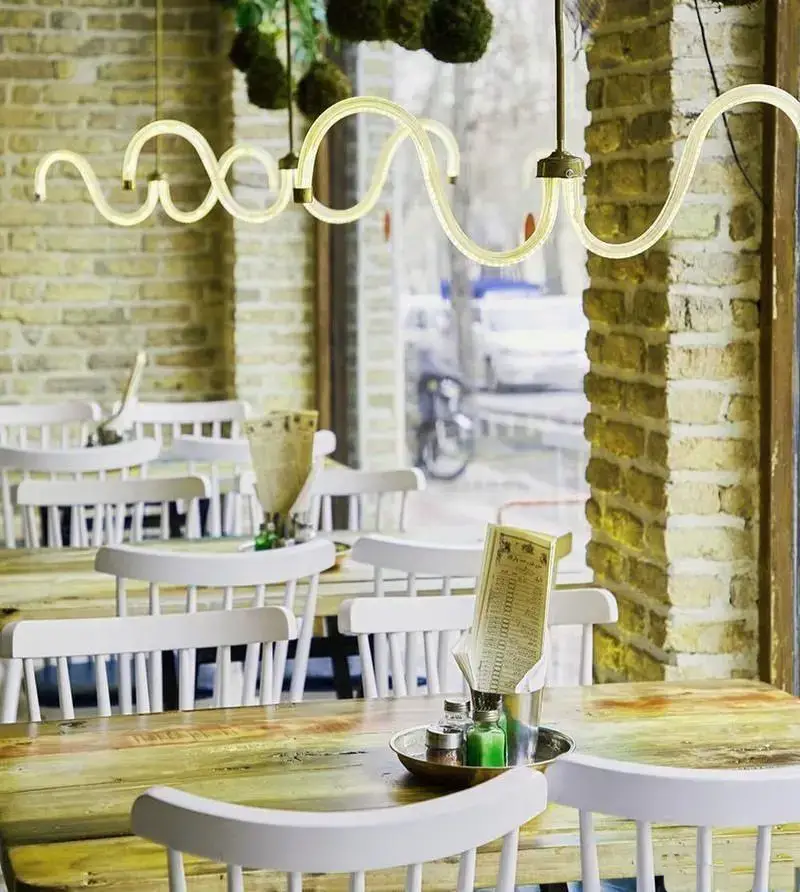 فضای داخلی رستوران ایرانی چارگاه با دکوراسیون گرم، میزهای چوبی و صندلی‌های سفید. لوستر موجی شکل نورپردازی خاصی به محیط بخشیده است.