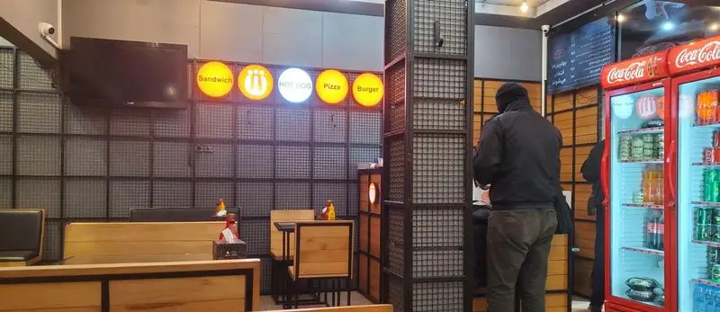 محیط داخلی رستوران فست فود زنجیره ای شیلا با منوی نورانی ساندویچ، هات داگ، پیتزا و برگر، و یخچال‌های نوشیدنی کوکاکولا.