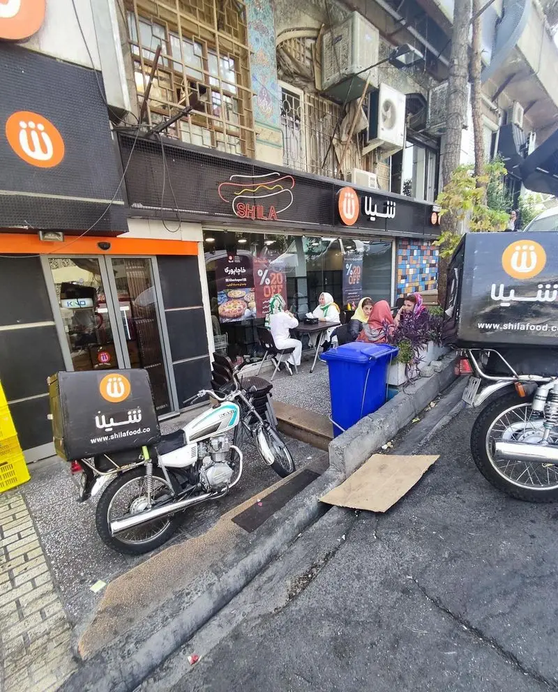 تصویری از نمای بیرونی شعبه فست فود زنجیره‌ای شیلا، با مشتریان نشسته، لوگو، تخفیف‌ها و پیک‌های موتوری آماده دلیوری غذا.