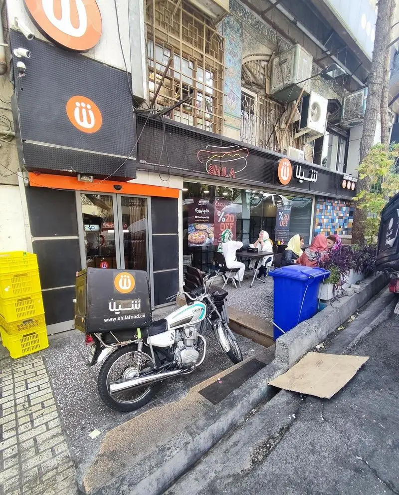 نمای شلوغ بیرونی رستوران فست فود زنجیره ای شیلا با لوگوی هات داگ برجسته، مشتریان و موتور سیکلت تحویل آنلاین غذای شیلا.