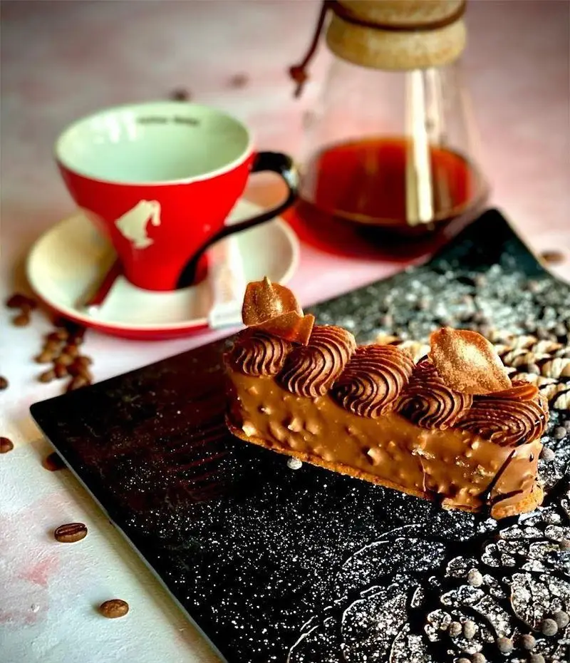 کیک شکلاتی خاص و خوشمزه کافه رستوران شاندیز گالریا با تزیین خامه و مغز، در کنار فنجان قهوه و قهوه دَم شده تازه.