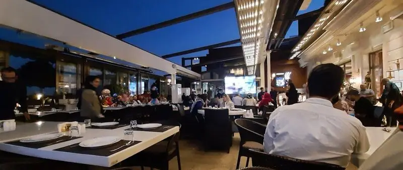 کافه رستوران شاندیز گالریا در شب با مشتریان در حال صرف غذا و نوشیدنی. فضایی باز با سقف متحرک و نورپردازی زیبا، ایده‌آل برای دورهمی‌ها.