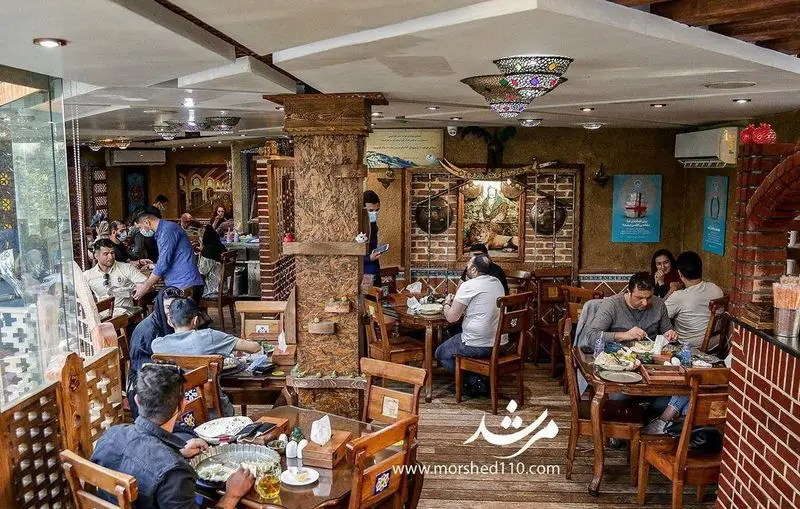 نمایی از فضای داخلی رستوران سنتی مرشد باهنر پر از مشتریانی که غذا می خورند و محیطی دلنشین با دکوراسیون چوبی و آجری.
