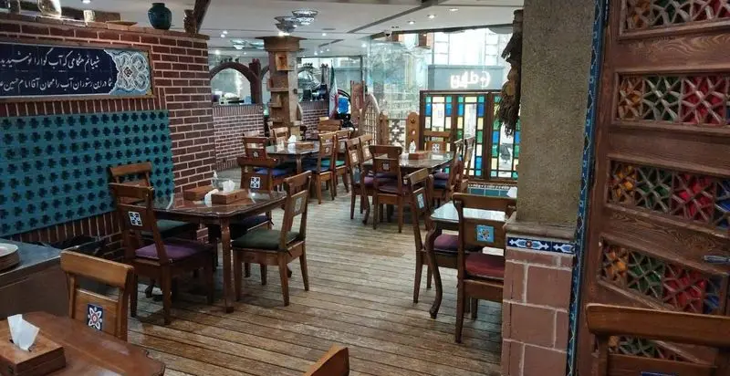 فضای داخلی دلنشین و سنتی رستوران مرشد باهنر با میز و صندلی‌های چوبی، دیوار آجری، کاشی‌کاری‌های آبی و شیشه‌های رنگی.